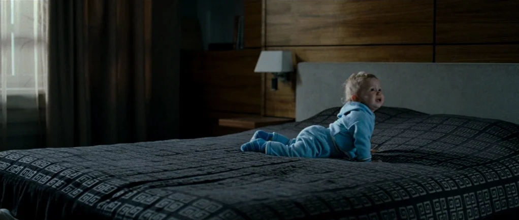 Elena (2011) movie still: wide — A modern bedroom features a large bed with a dark, patterned cover and headboard. A small child in a blue suit…