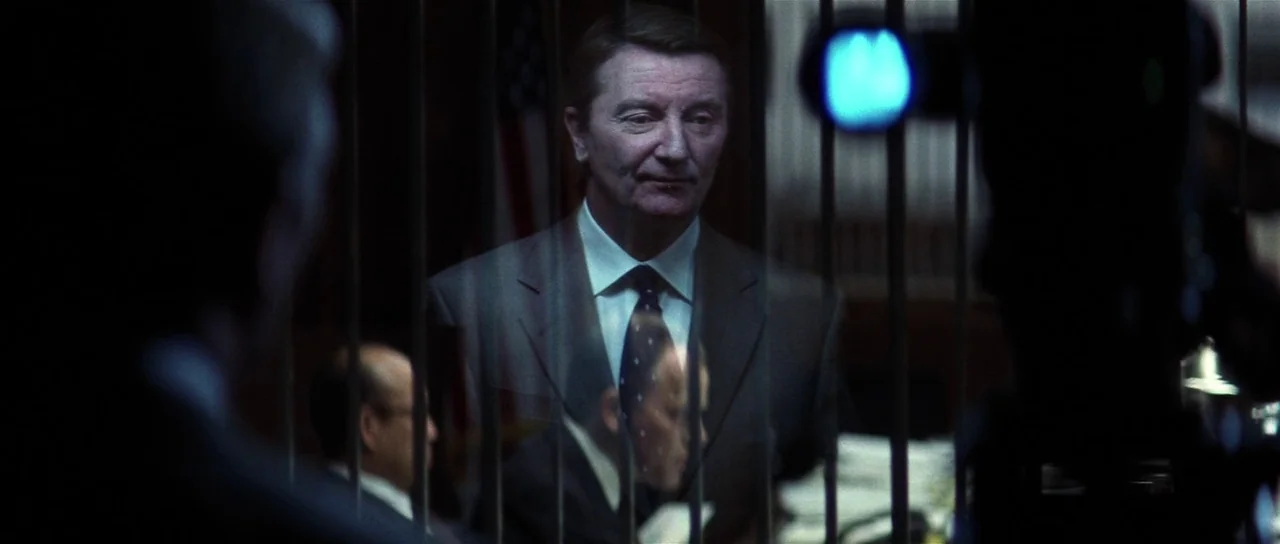 Spy Game (2001) movie still: medium — A man stands behind a vertical metal grate in a dimly lit office. Behind him hangs a blurred American flag…