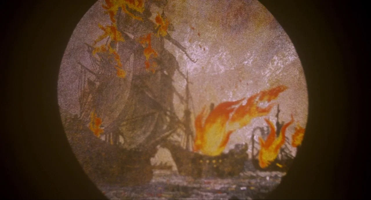 Ammonite (2020) movie still: establishing — A circular view shows a naval battle with wooden ships engulfed in bright flames. The ships are…