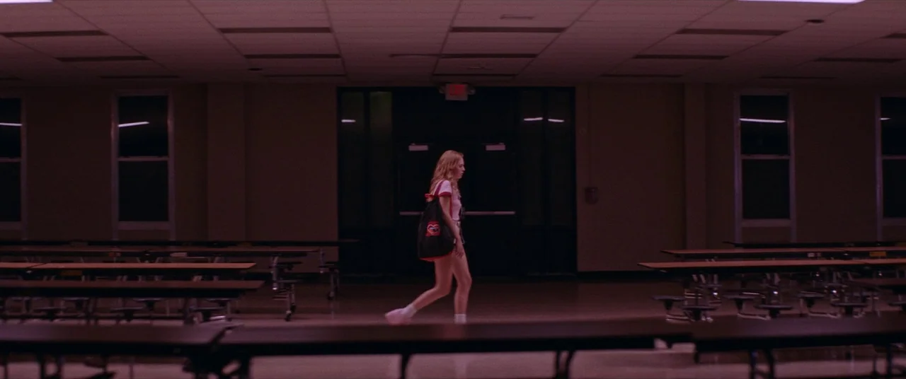 Assassination Nation (2018) movie still: wide — An empty school cafeteria features rows of long tables with attached stools under dark lighting. A…