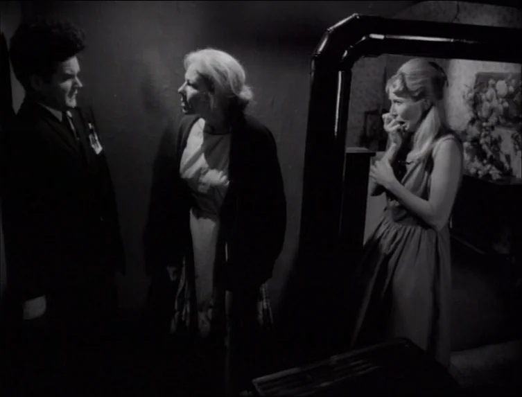 Eraserhead (1977) movie still: medium — A dark domestic interior with floral wallpaper and a heavy overhead ventilation pipe. Three people are…