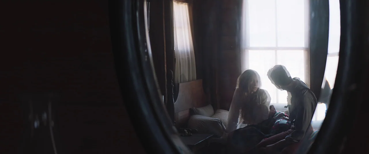 The Wind (2018) movie still: medium — A rustic log cabin bedroom features a wooden bed frame beside a large window. A man and a woman sit facing one…