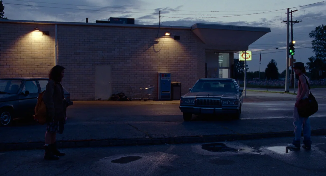 Bones and All (2022) movie still: wide — A quiet parking lot at a commercial building during twilight with a parked sedan. Two teenagers stand at…