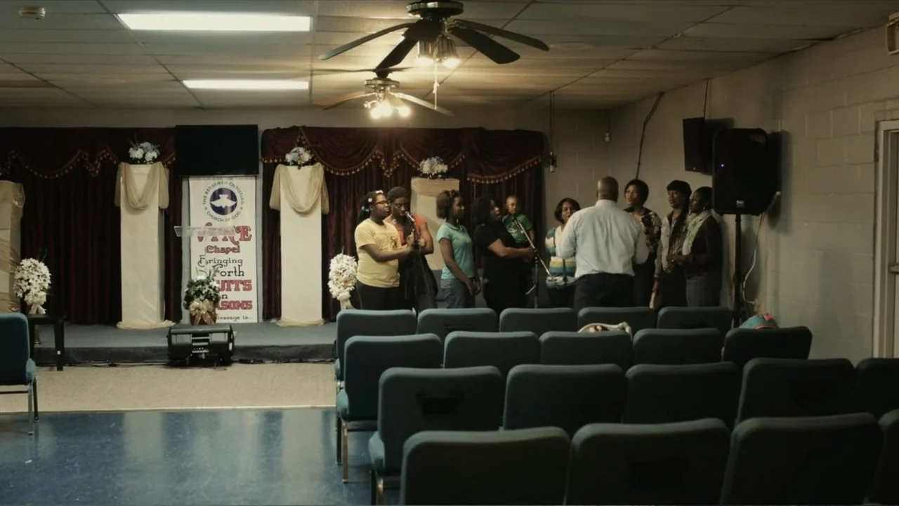Death of a Child (2017) movie still: wide — A small church sanctuary features blue upholstered chairs facing a stage with velvet curtains. A diverse…