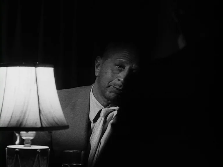 The Killing (1956) movie still: medium — A dimly lit interior space features a man positioned near a glowing table lamp. Deep shadows obscure the…