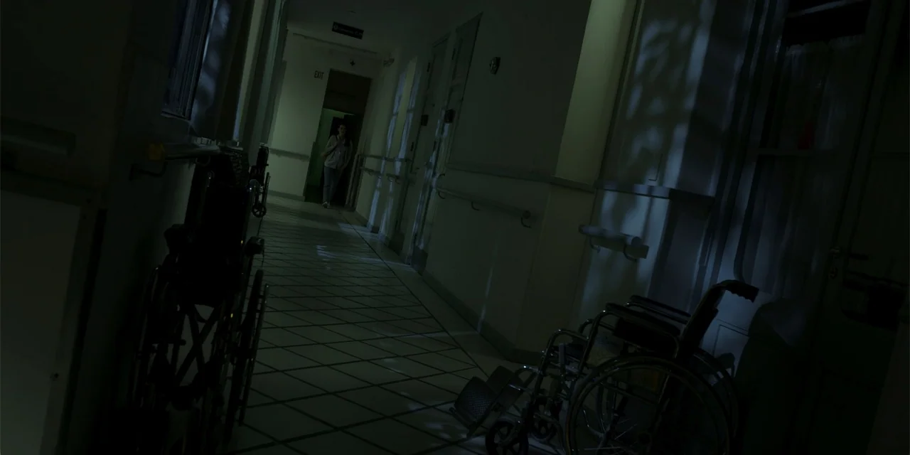 La coda del diavolo (2024) movie still: wide — A dimly lit hospital corridor features tiled flooring and two wheelchairs parked against the walls. A…