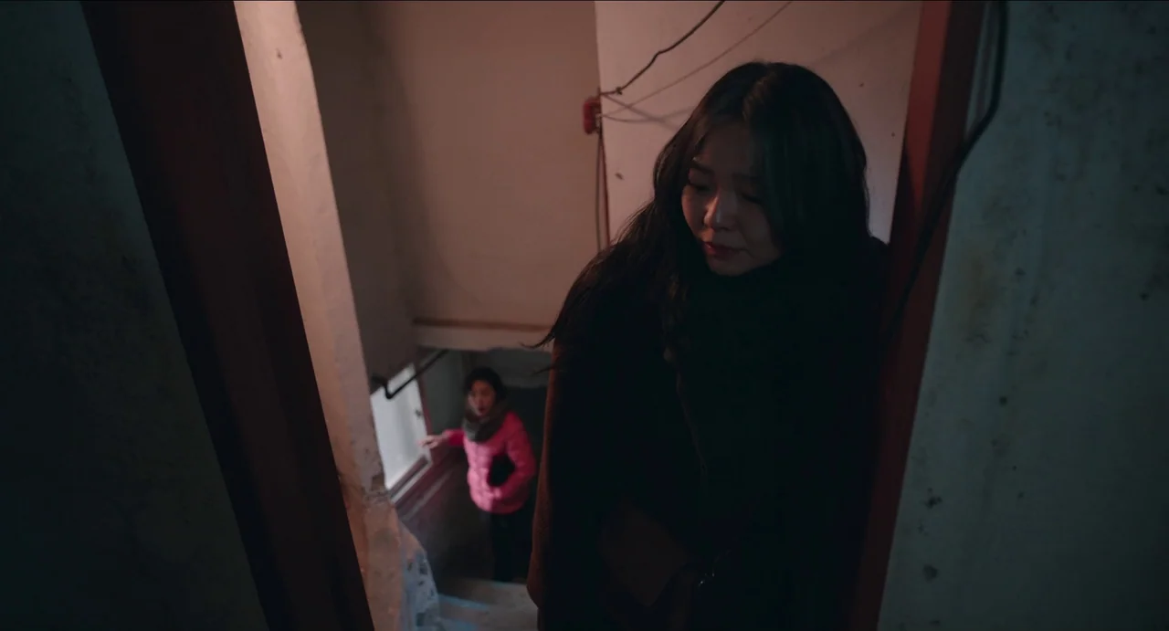 Microhabitat (2017) movie still: medium — An interior staircase with white walls and exposed electrical wiring overhead. A woman in a dark coat…