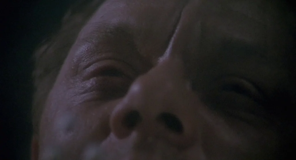 In a Glass Cage (1986) movie still: closeup — A dark, underexposed environment surrounds the upper half of a man's face. The subject is oriented…