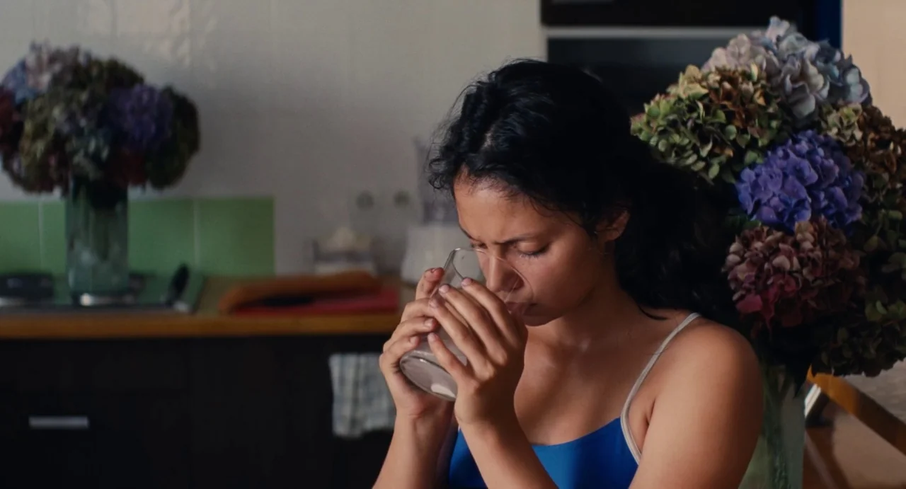 Ava (2017) movie still: medium — A woman stands in a kitchen with large bouquets of blue and purple hydrangeas. She holds a glass of water to her…