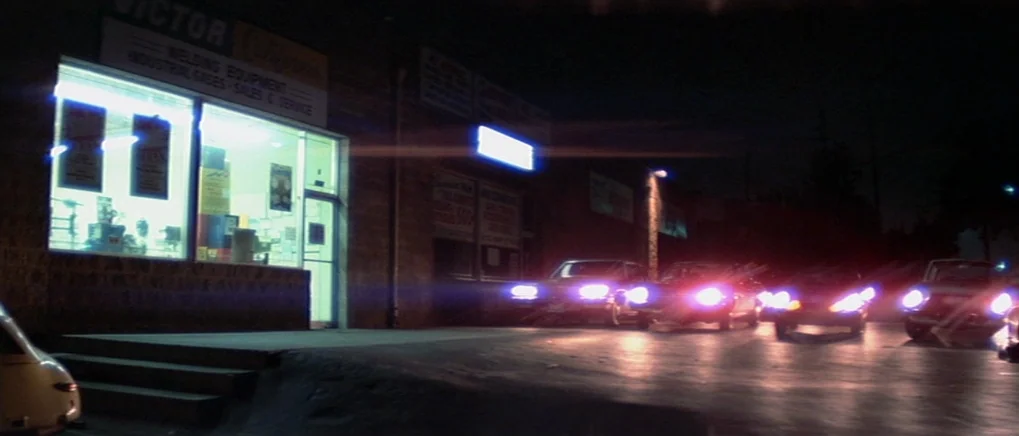 The Fog (1980) movie still: wide — A dark, low-slung industrial building features a brightly lit display window for welding supplies. Several cars…