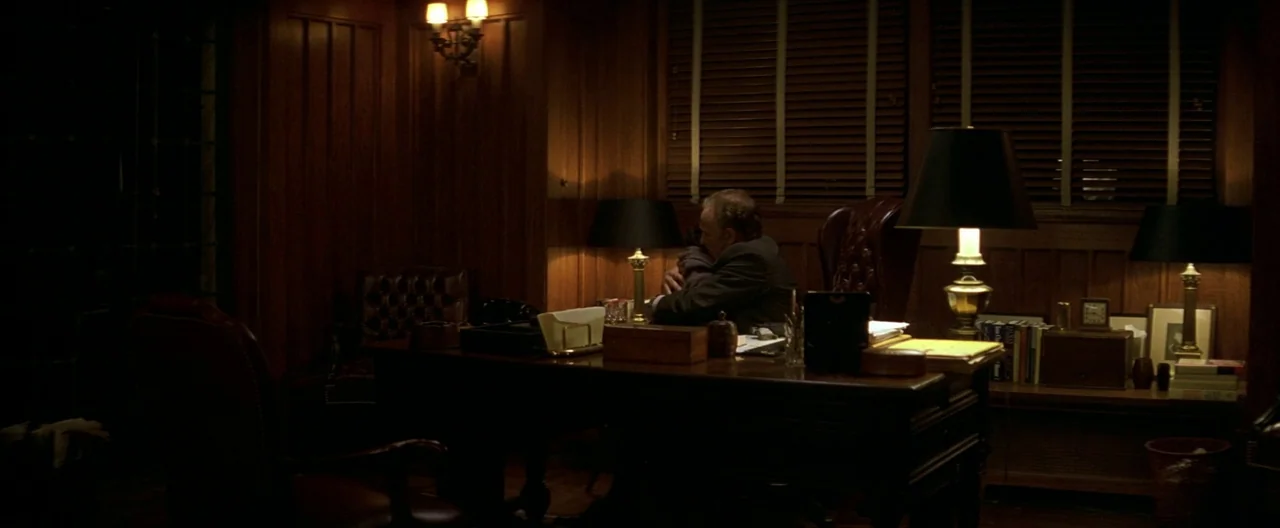 The Yards (2000) movie still: wide — A dark wood-paneled study features a large desk with multiple lamps and bookshelves. A man in a suit sits…
