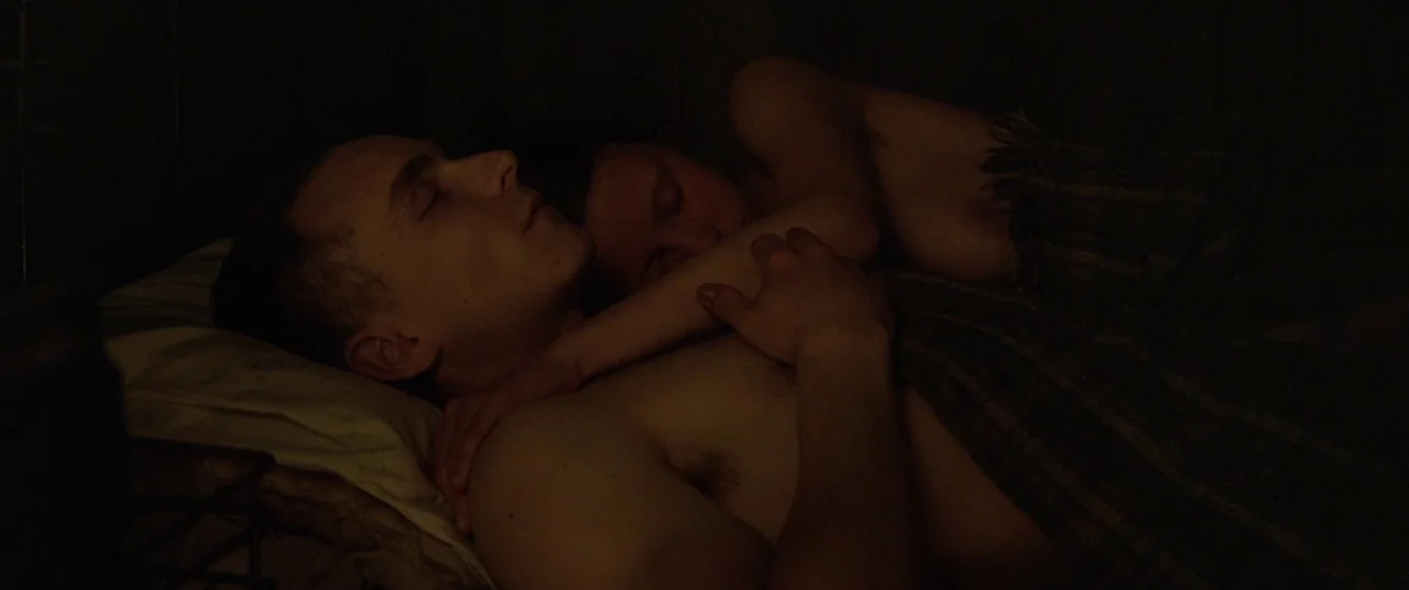 The Survivalist (2015) movie still: closeup — A dimly lit room with two people sleeping close together on a pillow. One person has their arm draped…