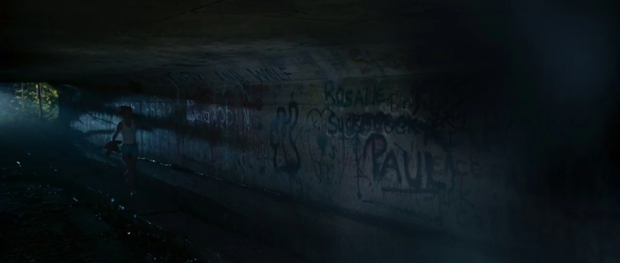 Silent Hill movie still: wide — A dark concrete underpass with extensive graffiti on the wall lit by distant light. A lone young woman runs through…