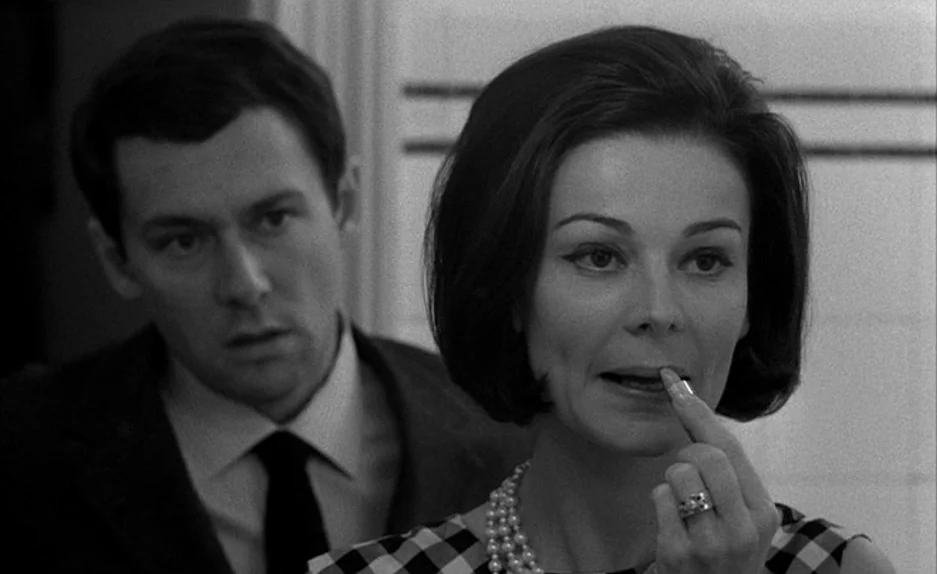 Le Feu Follet (1963) movie still: medium — A tiled room with a woman applying makeup reflected in a mirror. A man stands behind her looking toward…