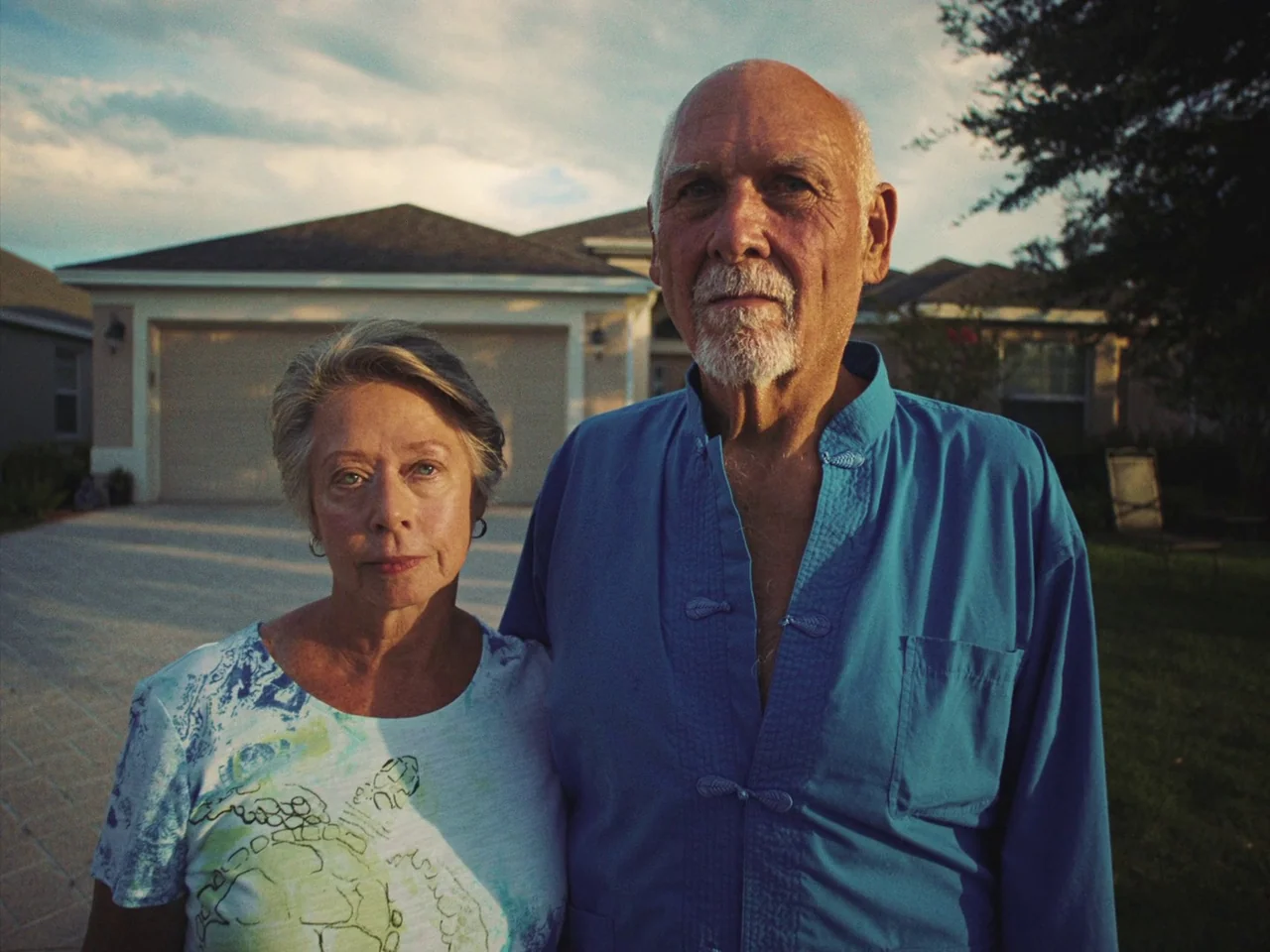 Some Kind Of Heaven (2020) movie still: medium — Two older people stand in a residential driveway during the late afternoon. A suburban house with a…