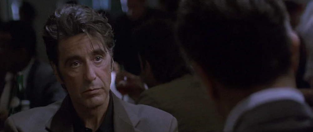 Heat (1995) movie still: closeup — A dimly lit indoor space with blurred patrons in the background. A man in a suit looks toward his conversational…
