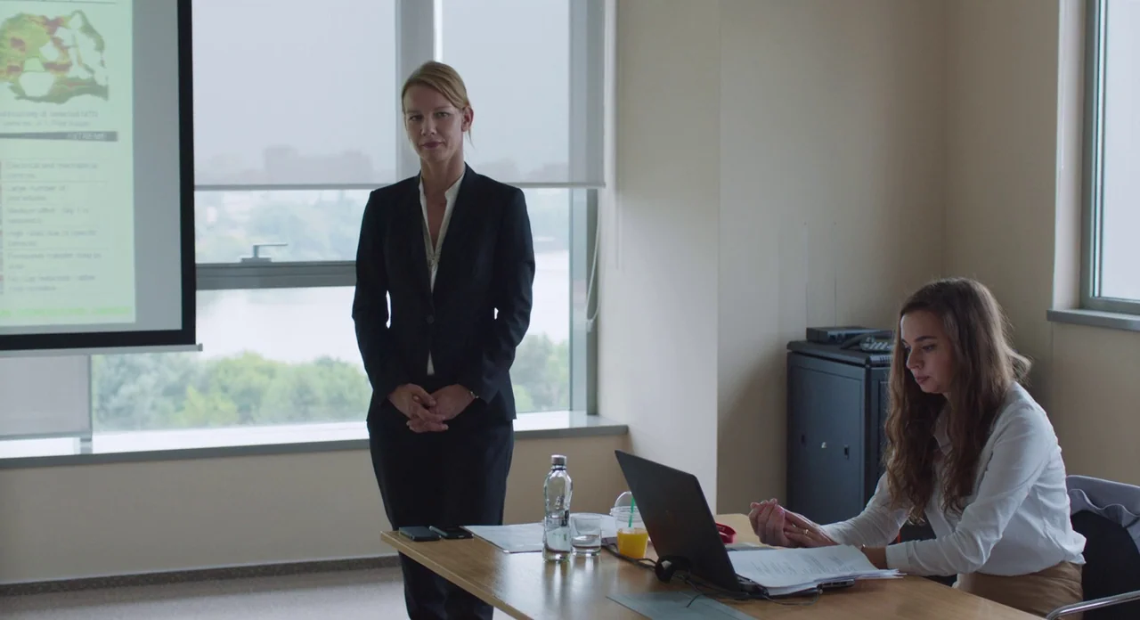 Toni Erdmann (2016) movie still: fullbody — A modern meeting room features a large window with white roller shades and monitors. A woman stands in…