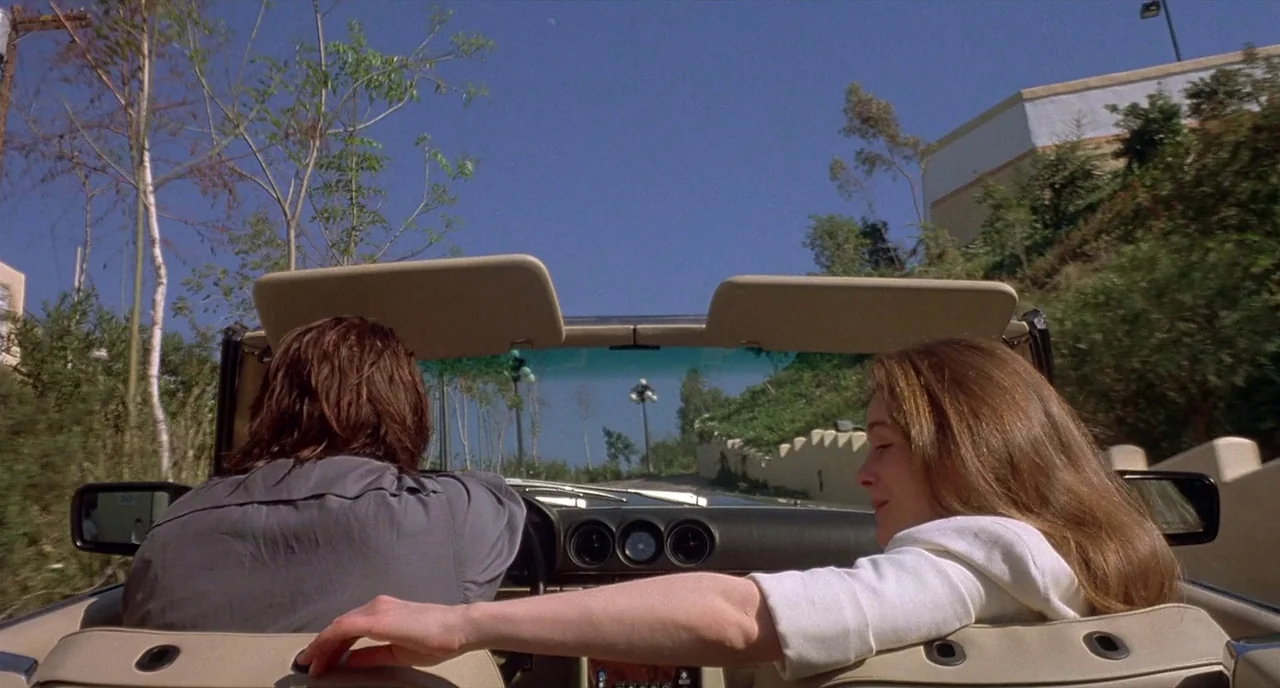 Barfly (1987) movie still: medium — Two people ride in a light-colored convertible through a sunny, hilly neighborhood. The man drives while the…