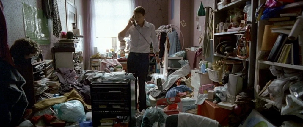 Next Door (2005) movie still: fullbody — A crowded, disorganized interior filled with piled clothes, books, and miscellaneous household objects. A…