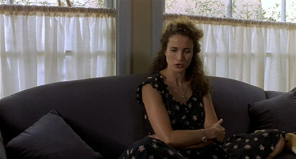 Sex, Lies And Videotape movie still: medium — A dark sofa sits in front of a pair of windows with translucent curtains. A woman with dark curly hair…