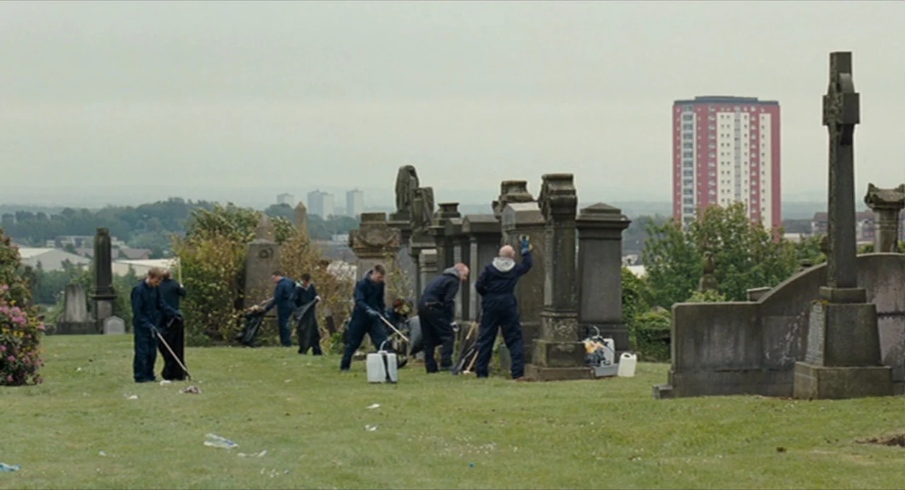 The Angel’s Share (2012) movie still: wide — A grassy cemetery with stone monuments overlooks a distant red apartment building. Several workers in…