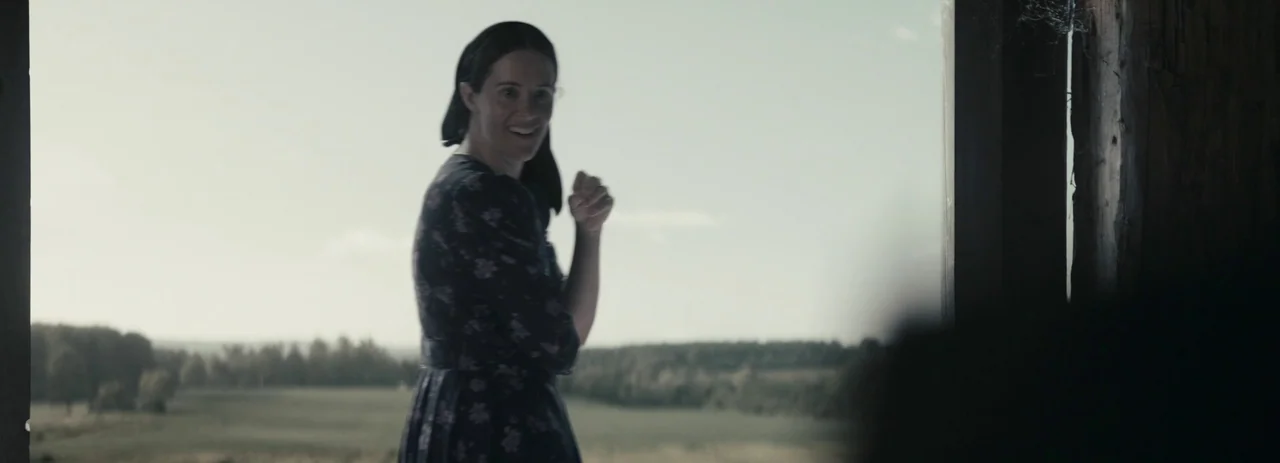 Women Talking (2022) movie still: medium — A woman stands in the shadows of a weathered wooden barn doorway. She gazes outward toward a vast,…