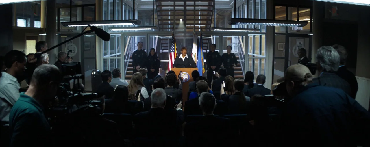 The Naked Gun (2025) movie still: wide — A spacious office interior features a glass-walled atrium with stairs in the background. A woman delivers a…
