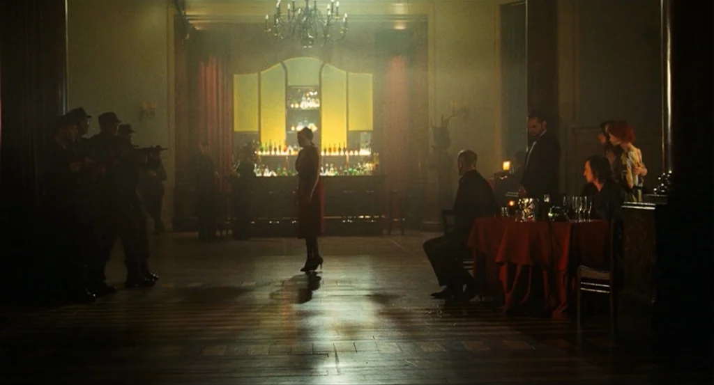This Night (2008) movie still: wide — A dark, wood-paneled ballroom features a central bar and scattered tables with red cloths. A woman stands in…