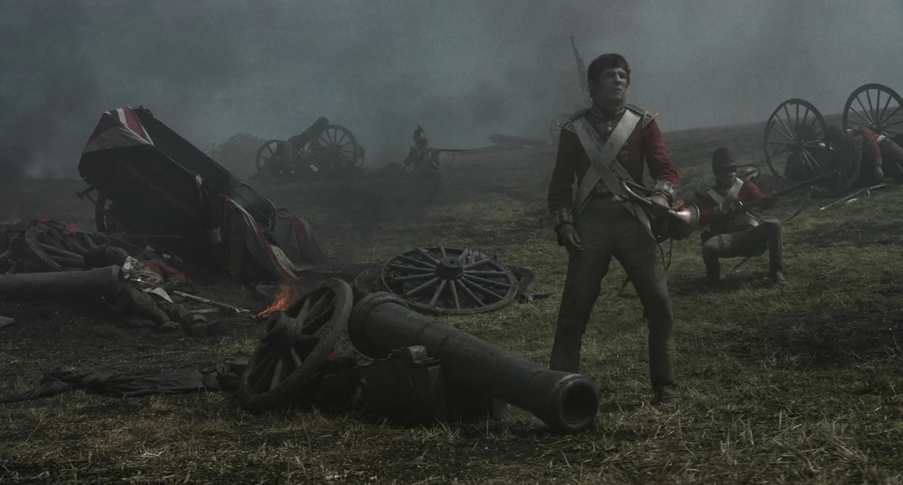 Peterloo (2018) movie still: fullbody — A smoke-filled 19th-century battlefield is covered in broken cannons and discarded carriage wheels. A young…