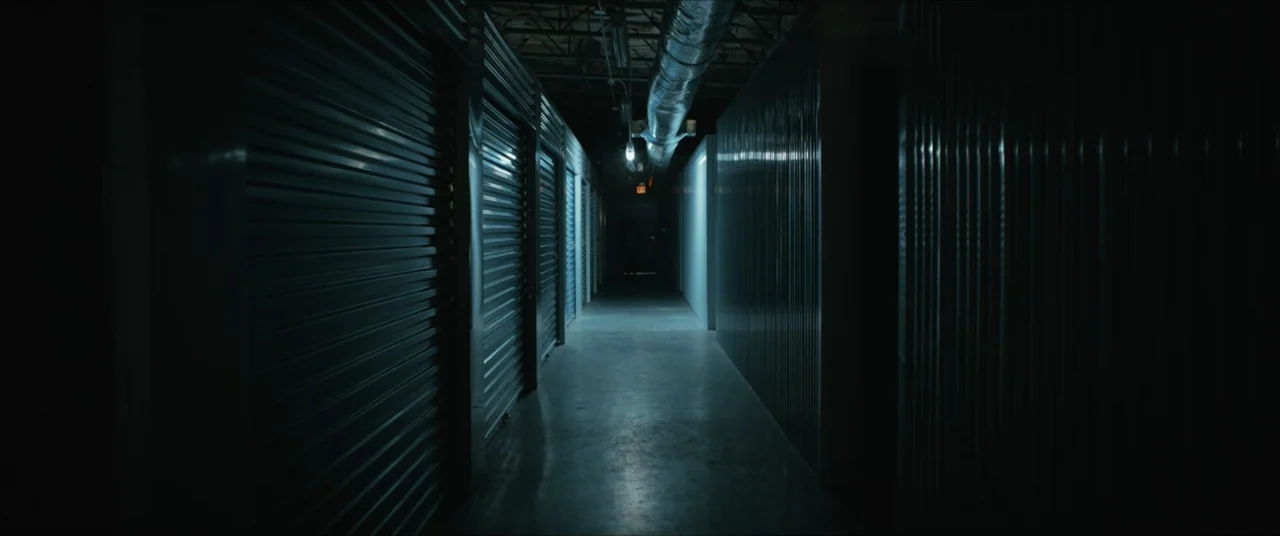 Possessions (2024) movie still: wide — A dark hallway of a storage facility lined with closed metal roll-up doors. The space is empty and…