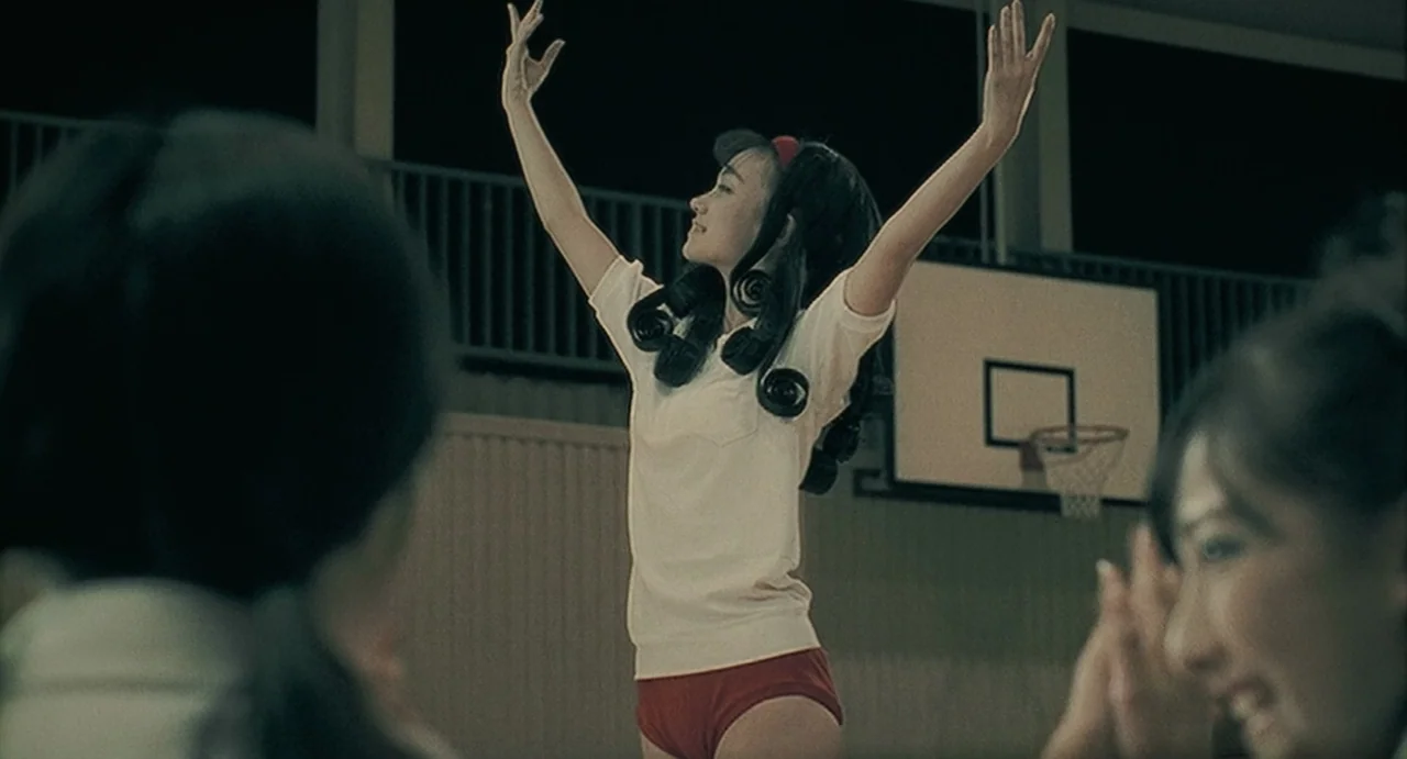 Uzumaki (2000) movie still: medium — An indoor school gymnasium features a basketball hoop mounted on the rear wall. A teenage girl in athletic…