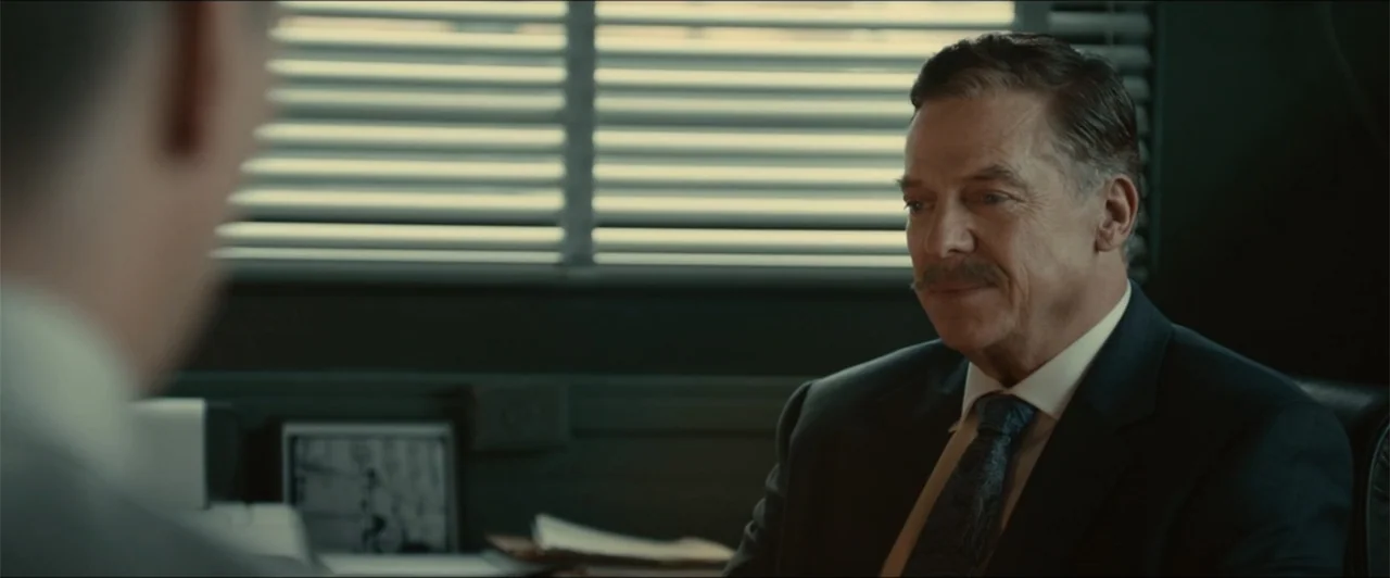 Exposed (2016) movie still: medium — A dimly lit office features horizontal window blinds filtering daylight into the room. An older man in a suit…