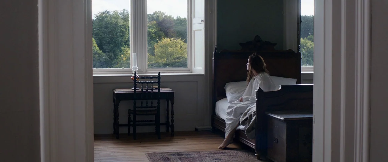 Lady Macbeth (2016) movie still: wide — A spare, wood-floored room features a high-backed dark wooden bed frame and desk. A woman sits on the edge…