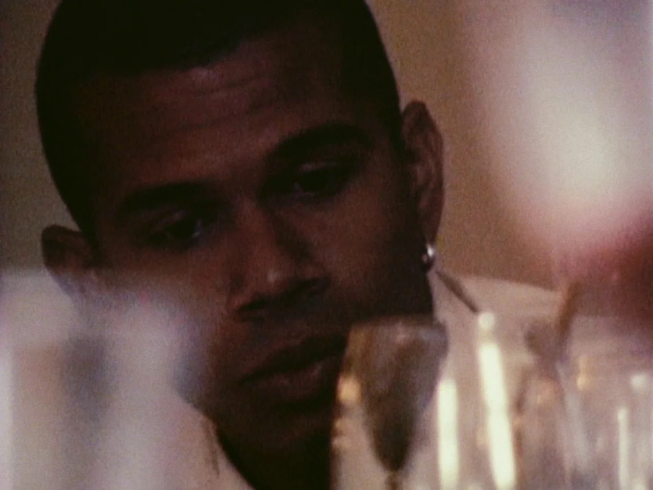 Festen (1998) movie still: closeup — A man sits at a table in a dimly lit, warm-toned dining space. He looks down at a glass while the foreground is…
