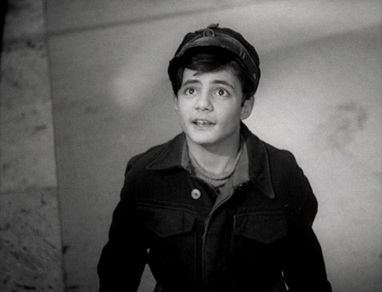 I Vitelloni (1953) movie still: medium — An interior stone stairwell with smooth walls and a textured floor surface. A young boy wearing a cap looks…