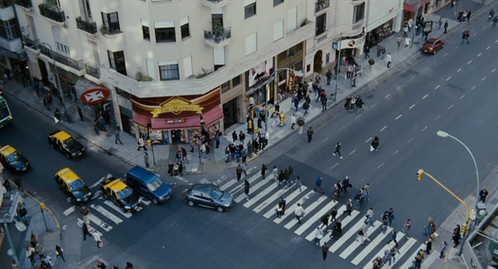 Sidewalls (2011) movie still: establishing — A high-angle view of a busy city corner with a crosswalk and traffic. Numerous pedestrians walk through…