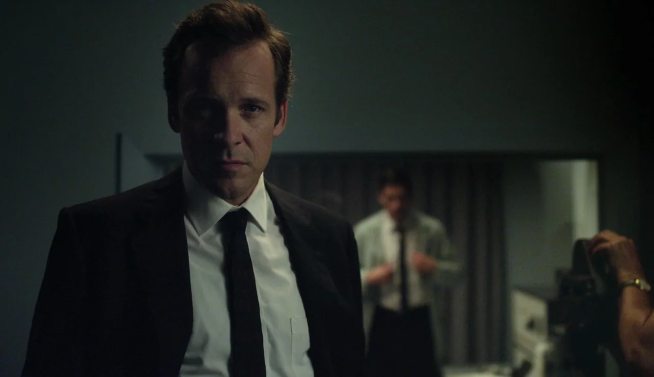 Experimenter (2015) movie still: medium — A dimly lit, sparse interior space features a man in a formal suit. In the blurred background, another…