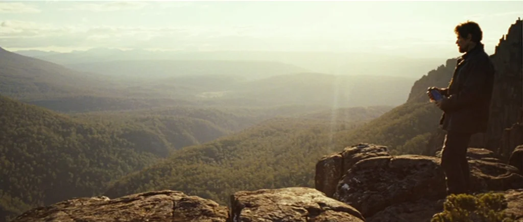 The Hunter (2011) movie still: wide — A vast mountain range stretches into the distance under a hazy golden sky. A man stands on the rocky edge…