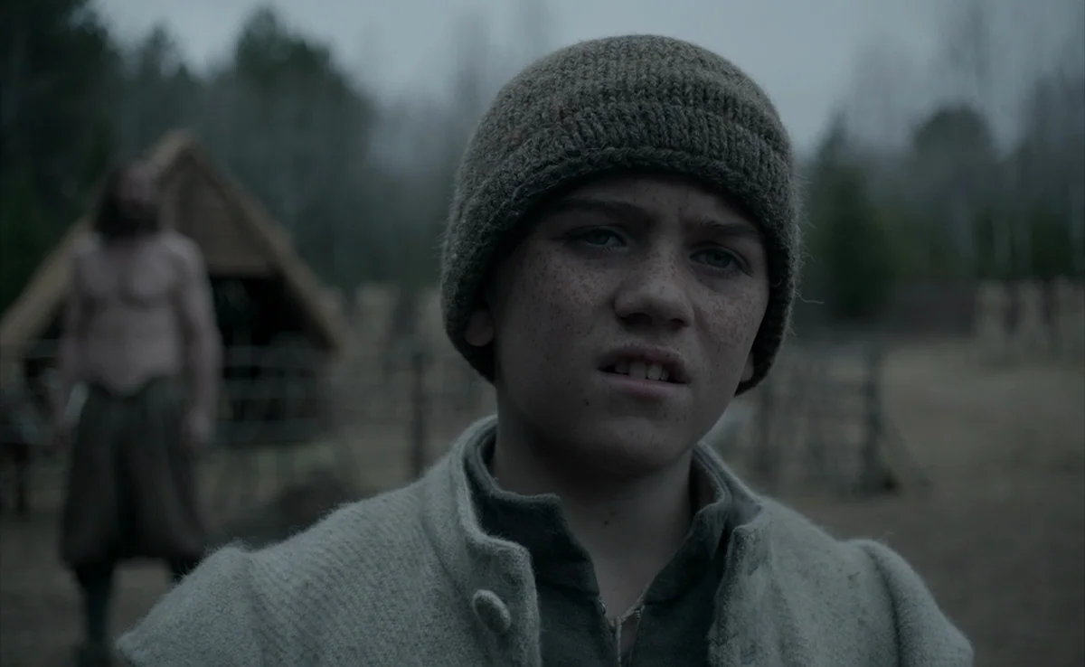 The Witch (2015) movie still: closeup — A rural, overcast outdoor setting features a rustic, wood-framed structure in the background. A young boy in…