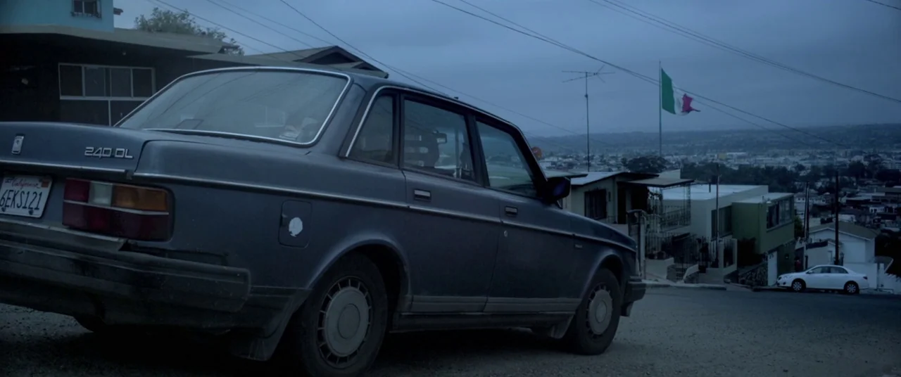 Here Comes The Devil (2012) movie still: wide — A dark blue Volvo sedan is parked on a downward sloping residential street. Houses line the hill…