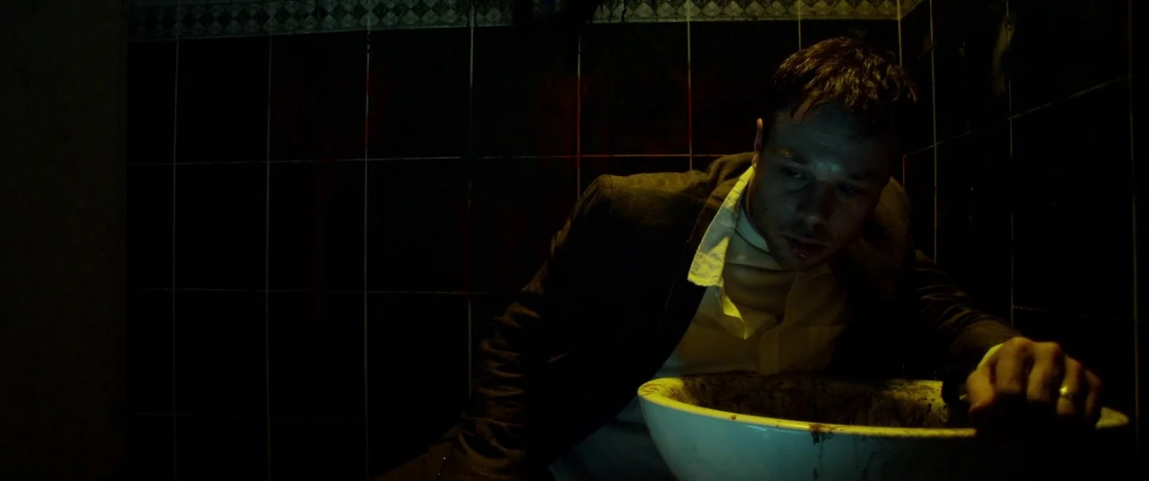 The Canal (2014) movie still: medium — A dark, tiled room features black wall tiles and a white porcelain basin. A disheveled man leans over the…