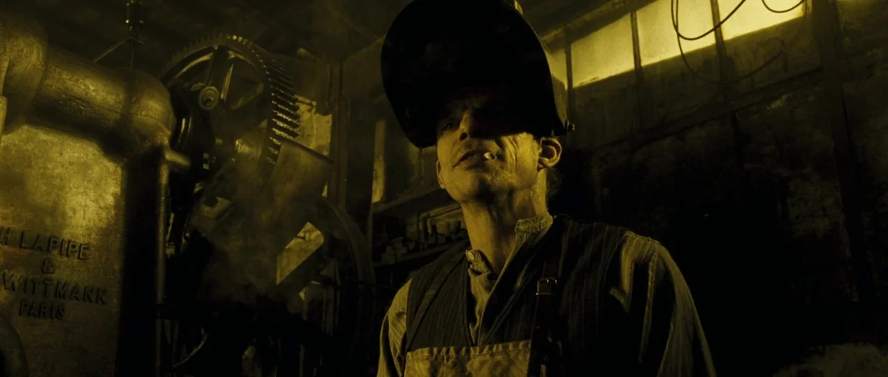 A Very Long Engagement (2004) movie still: medium — A dark industrial room filled with heavy metal machinery and gear mechanisms. A man wearing a…