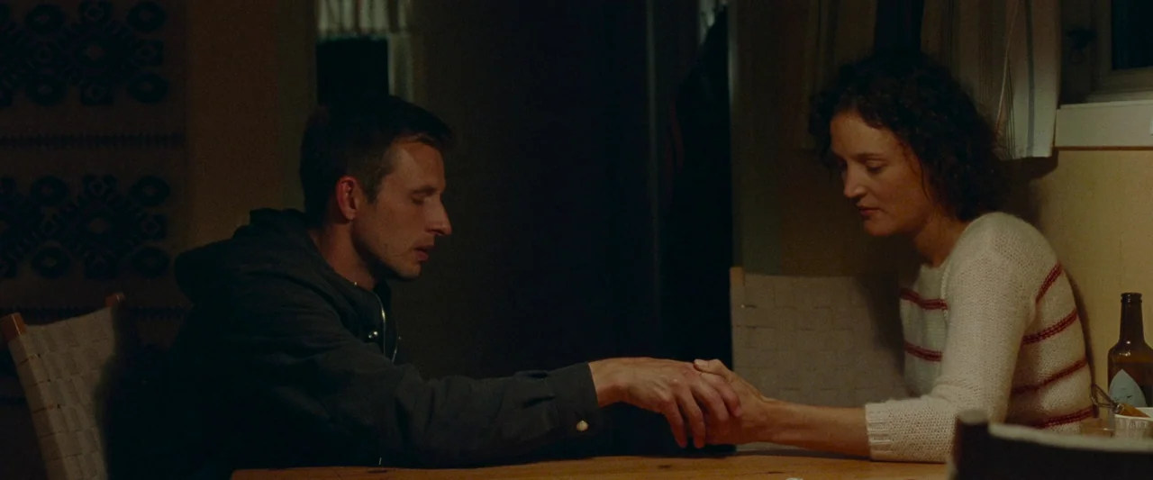 Bergman Island (2021) movie still: medium — A dimly lit room with a wooden table serves as the primary setting. A man and a woman sit facing each…