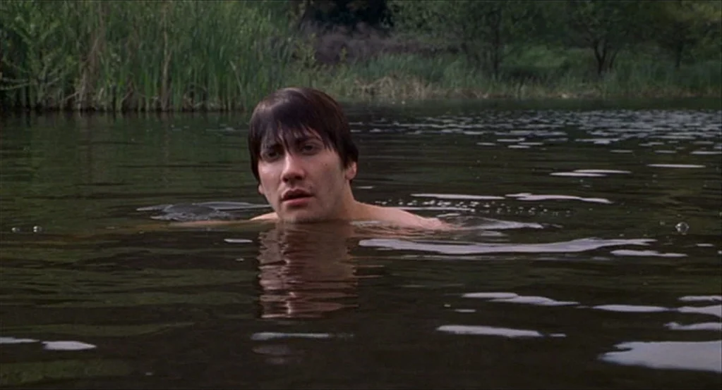 The Good Girl (2002) movie still: medium — A man floats in the dark, still water of a remote natural lake. Tall green reeds and distant trees line…