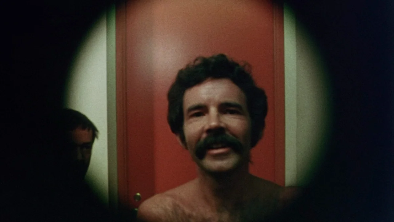 Shivers (1975) movie still: closeup — A man stands before a red door inside a room with heavy vignetting. A second person is partially visible in…