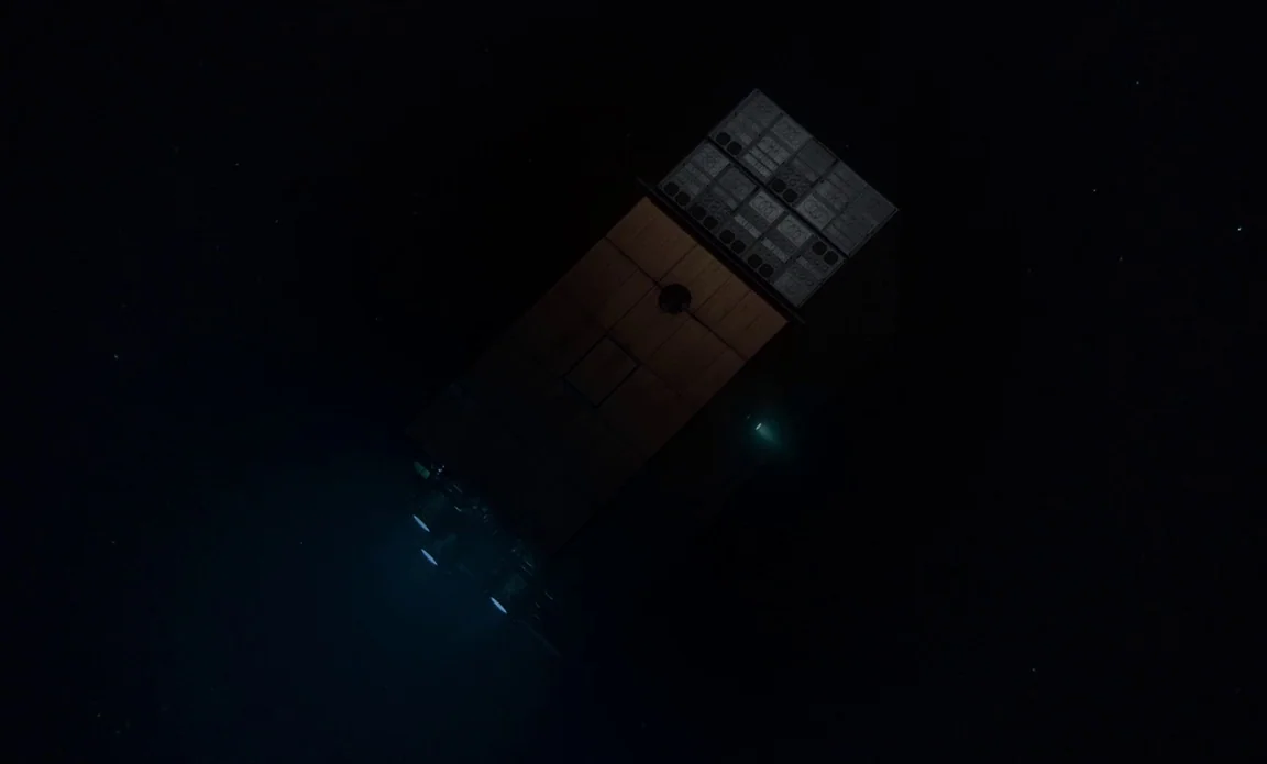High Life (2018) movie still: establishing — A rectangular spacecraft drifts through the dark void of deep space. Faint thruster emissions emit a…