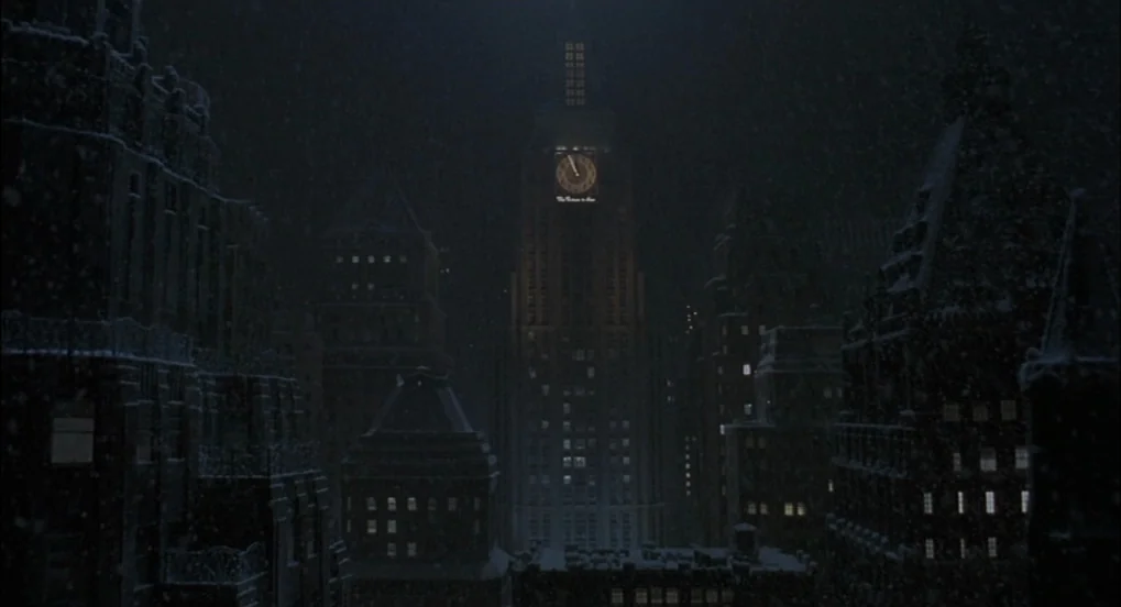 The Hudsucker Proxy (1994) movie still: establishing — A dense urban skyline at night features tall stone buildings covered in light snow. A central…