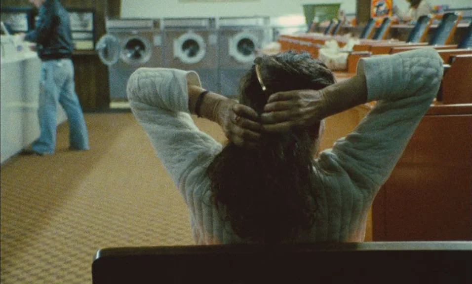 Documenteur (1981) movie still: medium — A 1970s laundromat with rows of brown machines and tan carpeting. A woman in a white sweater sits in the…