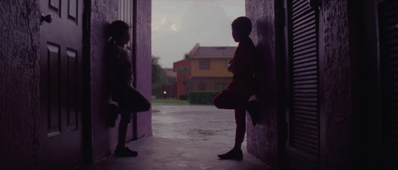 The Florida Project (2017) movie still: fullbody — A dark, purple-toned apartment breezeway frames a view of a suburban neighborhood during rain.…