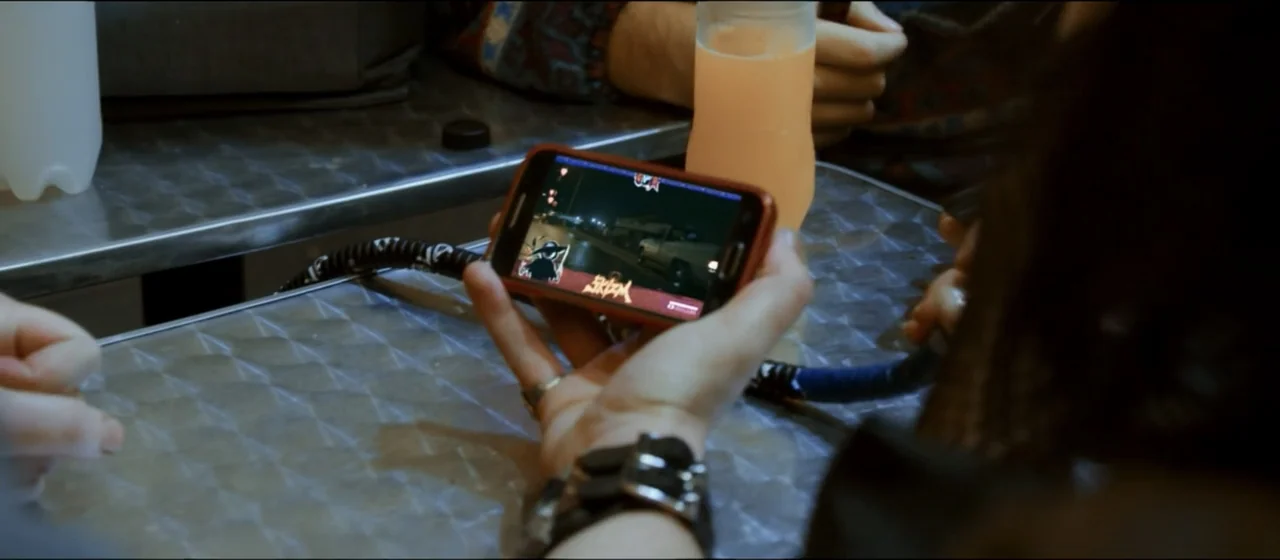 Guns Akimbo (2020) movie still: closeup — A person holds a smartphone over a patterned metallic table in a dimly lit setting. The screen displays a…